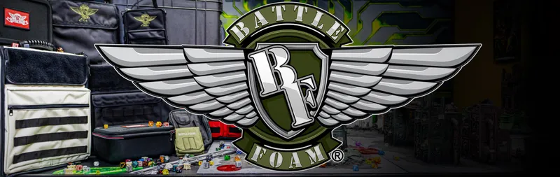 Stylized emblem featuring a shield design. At the top of the shield, bold white capital letters spell "BATTLE". In the center, a large white number "3" is integrated with a sword pointing downward. Below the shield, a green banner with white capital letters reads "FOAM" and to the right of it, a registered trademark symbol "(R)". The background depicts a cluttered tabletop with miniature terrain pieces, small figures, dice, and a red vehicle model. The setting appears to be a gaming or hobby workspace.