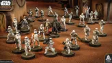A group of Star Wars miniatures features several Stormtroopers in white armor, positioned in a desert-like environment with a backdrop of a weathered building. Among them stands a prominent figure in black armor, holding a red lightsaber, likely representing Darth Vader. The scene is detailed with various poses of the troopers, some aiming weapons, while others appear to be in action, all set on circular bases with textured surfaces.