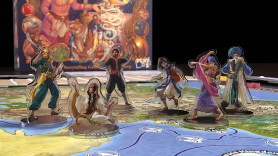 Five character standees are positioned on a colorful game board depicting a map inspired by Arabian Nights. The characters include a seated figure in white robes, a man in a red vest and black pants, a woman in a flowing pink outfit, a man in a blue vest with an orange satchel, and an older man in a blue turban and white robe. In the background, the game box is partially visible, featuring vibrant artwork and the text 