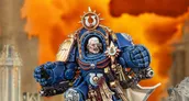 A detailed miniature of Marneus Calgar, a prominent character from the Warhammer 40,000 universe, is depicted in a striking pose. He wears ornate blue power armor adorned with gold embellishments, featuring a prominent Ultramarines symbol on his chest and a skull motif. The background is a dramatic orange, suggesting a fiery battlefield, enhancing the intensity of the scene.