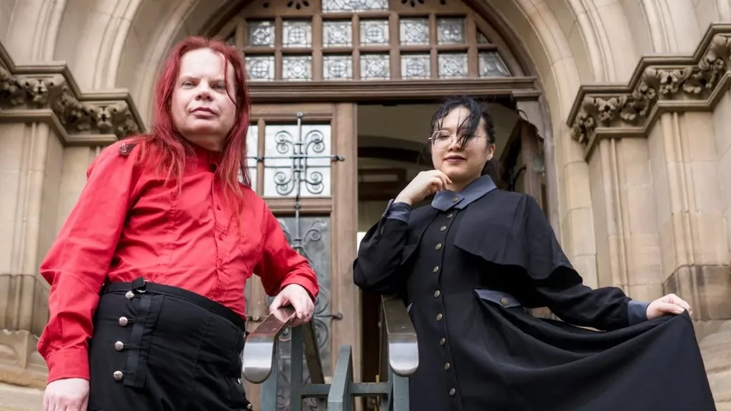 Two individuals pose in front of a grand, gothic-style building. One person, wearing a bright red shirt and black trousers with red stripes, stands confidently with arms crossed. The other person, dressed in a long black coat with a high collar and a blue shirt underneath, holds the hem of their coat, creating a dramatic silhouette. The ornate entrance of the building, featuring large wooden doors and intricate stonework, serves as a striking backdrop.