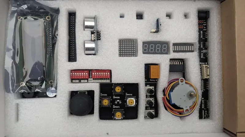 A collection of electronic components is neatly arranged on a white foam background. Key items include a pair of ultrasonic sensors, a digital display, various buttons, a relay, and a DIP switch. Additional components like a rotary encoder and several connectors are also present, showcasing a variety of parts typically used in DIY electronics projects.