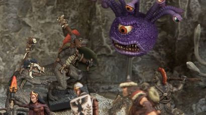 A collection of fantasy miniatures features several lizardmen, one wielding a shield and weapon, and another with a torch, set against a rocky backdrop. In the background, a large, purple creature with multiple eyes and a menacing expression looms, adding to the scene's fantastical atmosphere. A female character in dark robes stands nearby, while other figures in armor are partially visible, enhancing the sense of adventure.