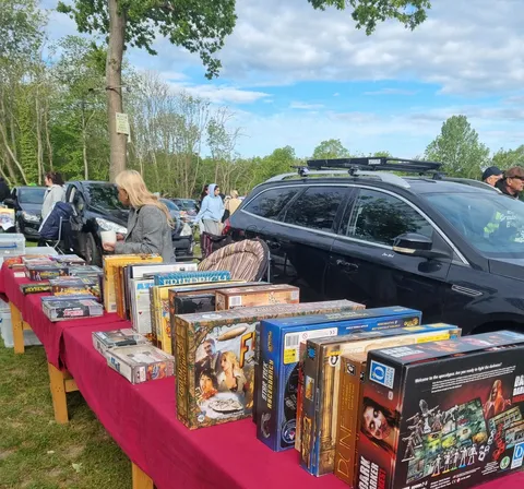 Outdoor car boot sale with red tablecloths displaying board games, DVDs, and books. People browsing and parked cars in background.