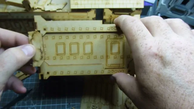 A hand holds a light green plastic model building piece, designed to resemble a fortified or industrial structure. The piece features four rectangular window cutouts arranged in two rows, each window framed with raised borders. Along the top edge, small circular holes are evenly spaced. The model appears to be part of a modular terrain set, resting on a patterned surface. The background shows additional similar model pieces, slightly out of focus. No text is visible on the model.
