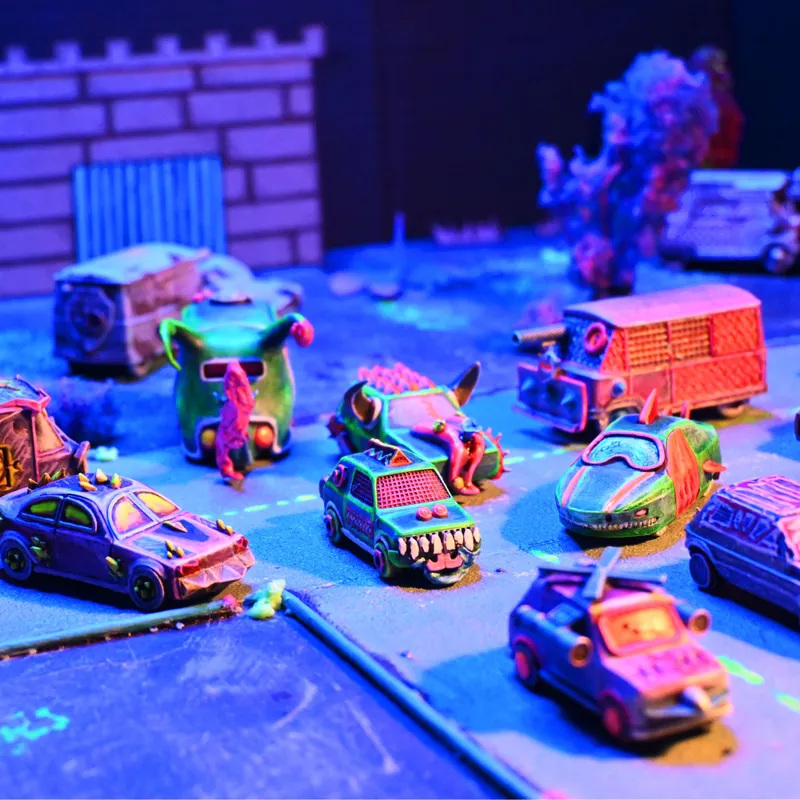 A vibrant tabletop scene features a variety of uniquely designed miniature vehicles, each with whimsical and chaotic elements, such as spikes, exaggerated features, and colorful paint jobs. The vehicles are arranged on a textured surface that resembles a racing track, with a backdrop of stylized scenery, including a stone wall and scattered debris. The lighting adds a dynamic flair, enhancing the fantastical atmosphere of the racing game "Fool's Fuel."