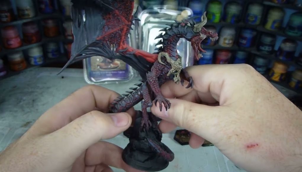 A detailed dragon miniature is being held in a hand, showcasing its intricate features, including prominent scales, fierce facial expression, and large wings with a red underside. The dragon is painted in dark hues with highlights that accentuate its muscular form and menacing appearance. In the background, various paint jars are visible, indicating a workspace for model painting.