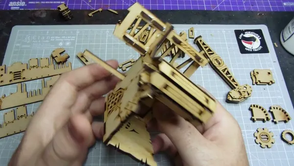 A partially assembled oil pump model made from laser-cut MDF is being held in hands, showcasing intricate details like vents and supports. Surrounding the model are various unassembled MDF pieces, indicating ongoing construction. A cutting mat with a grid pattern serves as the work surface, and a sticker reading "Orc Miniatures Hate Frogs" is visible in the background.