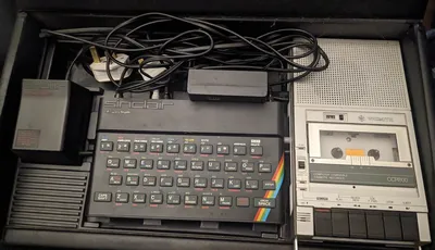 A Sinclair ZX Spectrum computer is prominently displayed, featuring a compact keyboard with colorful keys and a distinctive rainbow stripe along the side. Surrounding the computer are various cables and a power adapter, along with a cassette tape recorder positioned to the right. The scene is set within a case, suggesting a portable setup for retro computing.