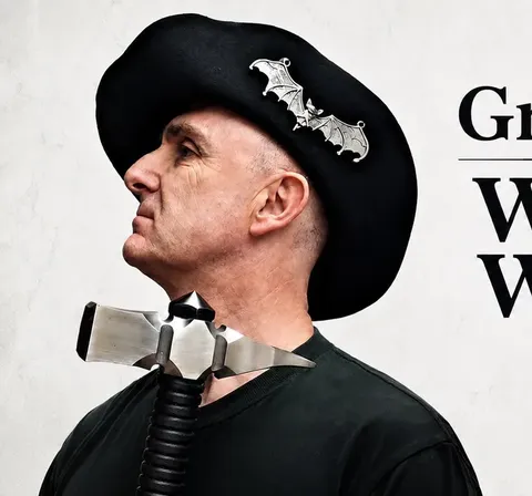 Man in black beret with bat pins, wearing a metallic microphone headset, black shirt.