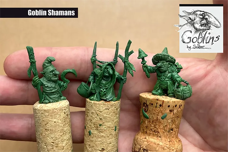 Three green goblin shaman miniatures are displayed on top of cork bases, showcasing their intricate details. The goblins feature various accessories, including staffs and bags, with distinct facial expressions that convey mischief. The background is a neutral color, allowing the vibrant green of the miniatures to stand out prominently.
