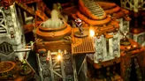 Miniature sci-fi figure in orange and white armor standing on a small platform atop an industrial terrain piece. Background shows metal pipes, grates, and a smokestack emitting vapor. Warm lighting highlights the scene.