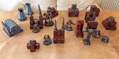 A collection of Epic scale terrain pieces suitable for games like Adeptus Titanicus and Battletech is displayed on a tan surface. The terrain includes various structures, such as a large, blue-gray building with pipes, several red industrial buildings, and tall smokestacks. Among the terrain, two detailed miniatures resembling mechs are positioned, adding to the scene's dynamic and futuristic atmosphere.