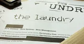 A close-up view of a printed document featuring the title "THE LAUNDRY" prominently displayed at the top, with a distressed font style. Below the title, the text indicates that it is based on the "LAUNDRY FILES" novels by Charles Stross, and mentions "System - New Management." The document appears to be part of promotional material for the latest edition of the Laundry RPG.