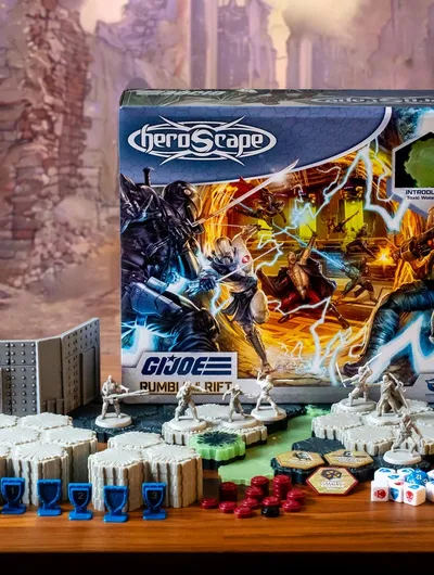 A G.I. JOE Heroscape: Rumble at the Rift Battle Box is prominently displayed in the center, featuring vibrant artwork of characters in action. Surrounding the box are various game components, including eight miniatures representing characters like Duke and COBRA Commander, modular hex terrain pieces, COBRA fortifications, dice, and markers. The background features a blurred, crumbling environment, enhancing the game's adventurous theme.