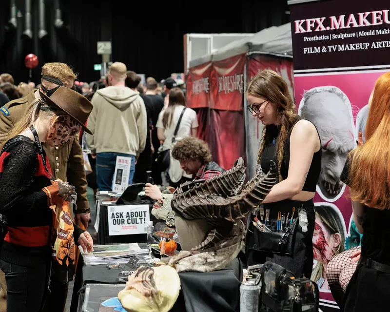 A bustling convention scene features attendees engaged in various activities, with a focus on horror themes. In the foreground, a woman dressed in a horror-themed outfit examines items on a table, while another attendee, wearing a brown hat, stands nearby. The table displays an array of props, including a large, intricate creature model, and promotional materials for horror-related events and products. The background is filled with other convention-goers and vendor booths, enhancing the immersive atmosphere of the event.