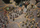Miniature dark fantasy wargame scene with two armies on a cracked, lava-like battlefield. Left side features dwarven figures with armor and weapons, including a large blue-robed figure with a glowing orb.