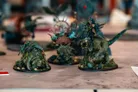 Three intricately painted tabletop miniatures are displayed on a flat surface. On the left, a large, armored creature resembling a tortoise is adorned with foliage and small details. In the center, a towering figure with a spiked shell and vibrant colors stands out, while on the right, a fierce, reptilian creature with a rider brandishing a weapon completes the trio. The background is slightly blurred, emphasizing the detailed craftsmanship of the miniatures.