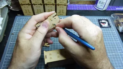 Two hands holding a partially assembled laser-cut MDF piece for a miniature building kit. The left hand supports the main structure, which is a yellowish-beige MDF panel with square cutouts and small circular holes. The right hand uses a blue-handled metal tool, likely tweezers or a small pick, to position or secure a small metal disc, possibly a magnet, into one of the circular holes. In the background, a clear plastic cutting mat with a printed grid is visible, along with several other laser-cut MDF sheets still in their frames, some with engraved or printed text and symbols. One of the MDF sheets in the background has a purple grid pattern. No readable text is visible on the main components being handled.