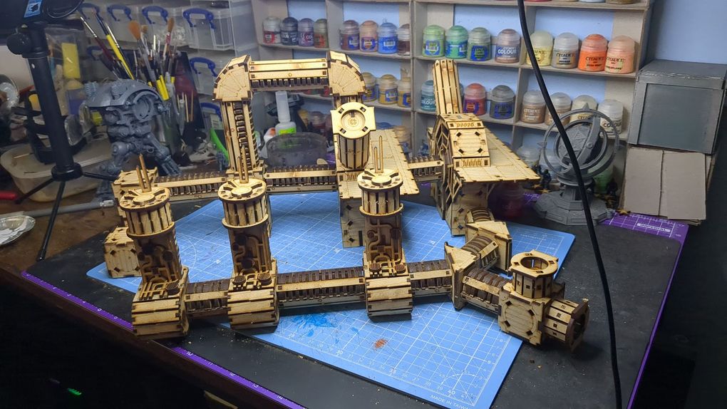 A detailed assembly of the TT Combat Sector 4 Plasma Relay is displayed, featuring intricate wooden structures with multiple towers and connecting pathways. The design includes cylindrical towers with circular tops and various platforms, all set against a blue cutting mat. In the background, jars of paint and tools are visible, indicating a workspace dedicated to tabletop gaming and model building.