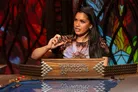 A woman with long black hair is animatedly speaking while gesturing with her right hand, seated at a table adorned with a "Dungeons & Dragons" sign. She wears a colorful shirt featuring a sun design and is positioned in front of a backdrop of stained glass, which adds a vibrant and atmospheric touch to the scene. The table is decorated with various gaming accessories, enhancing the tabletop roleplaying game setting.