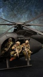 A detailed model of a Little Bird helicopter, featuring a dark color scheme, is prominently displayed. Two miniature soldiers are seated on the helicopter's skids, equipped with rifles, while another soldier is visible inside the cockpit. The background features a textured gradient, enhancing the focus on the helicopter and its crew.