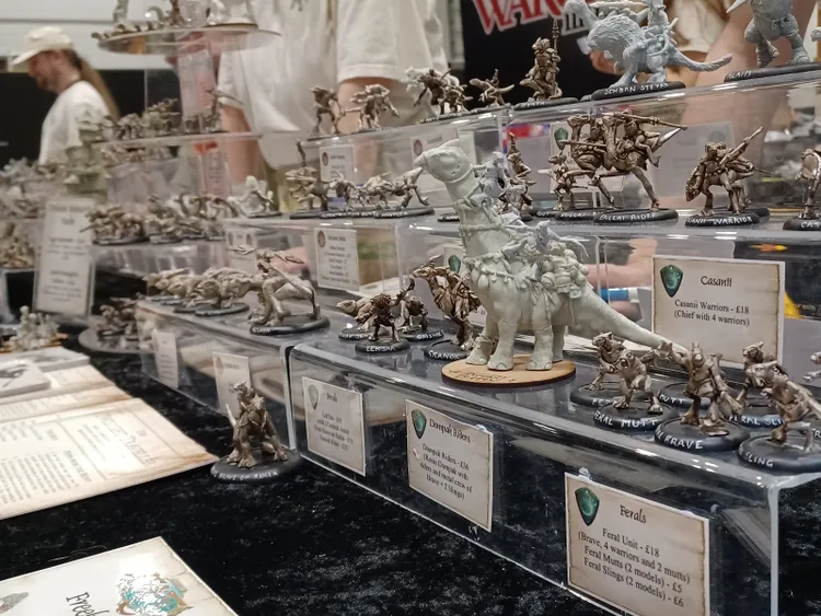 A display of various fantasy miniatures is showcased on a clear stand, featuring a mix of intricately detailed figures. Prominent in the center is a large, pale-colored creature resembling a dinosaur, with a rider atop, surrounded by smaller figures including warriors and anthropomorphic animals. Each miniature is labeled with its name and price, highlighting a diverse range of characters such as Casanii Warriors, Dumplok Riders, and Ferals, all set against a dark backdrop that enhances their features.