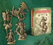 A collection of intricately designed miniatures from the Dragonbane RPG is displayed against a textured green background. The set includes a variety of characters, such as a knight with a spear, a dragon, and a sorcerer, all rendered in a beige color, showcasing their detailed features. To the right, a box labeled "DRAGONBANE DEMONS SET" features artwork of a menacing creature, indicating the theme of the miniatures within the set.