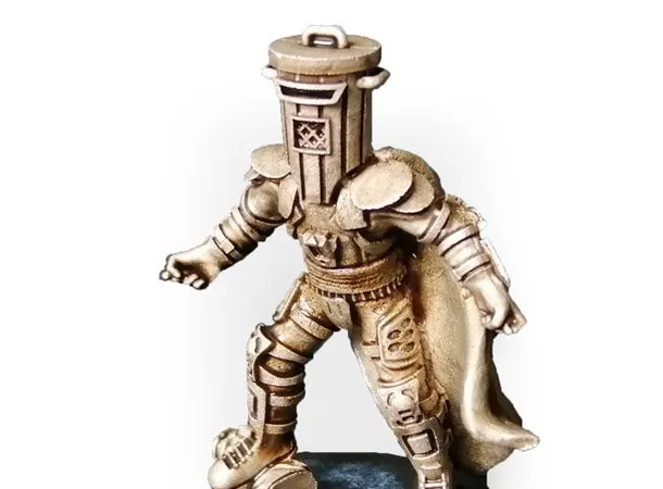 A gold-colored miniature figurine of a character wearing a cylindrical helmet resembling a trash bin, with a cape flowing behind. The figure stands on a round black base with a textured surface. The helmet has a handle on top and a small emblem on the front. The figure is posed with one arm extended forward and one leg slightly bent, as if mid-step.