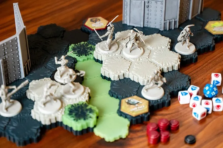 A tabletop scene features a modular hex terrain setup from the G.I. JOE Heroscape game, showcasing various hexagonal tiles in shades of green, black, and beige. Several miniatures, including characters wielding weapons, are positioned on the terrain, with some standing on raised hexes. Nearby, there are dice, wound markers, and Army cards, indicating the game's components for play.