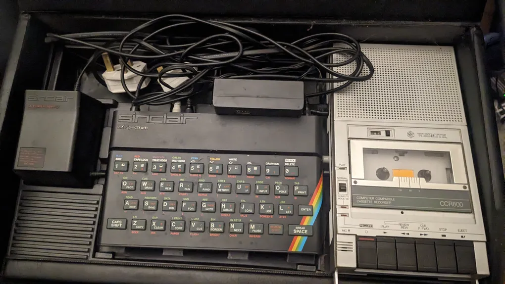 A Sinclair ZX Spectrum computer is prominently displayed, featuring a compact keyboard with colorful keys and a distinctive rainbow stripe along the side. Surrounding the computer are various cables and a power adapter, along with a cassette tape recorder positioned to the right. The scene is set within a case, suggesting a portable setup for retro computing.
