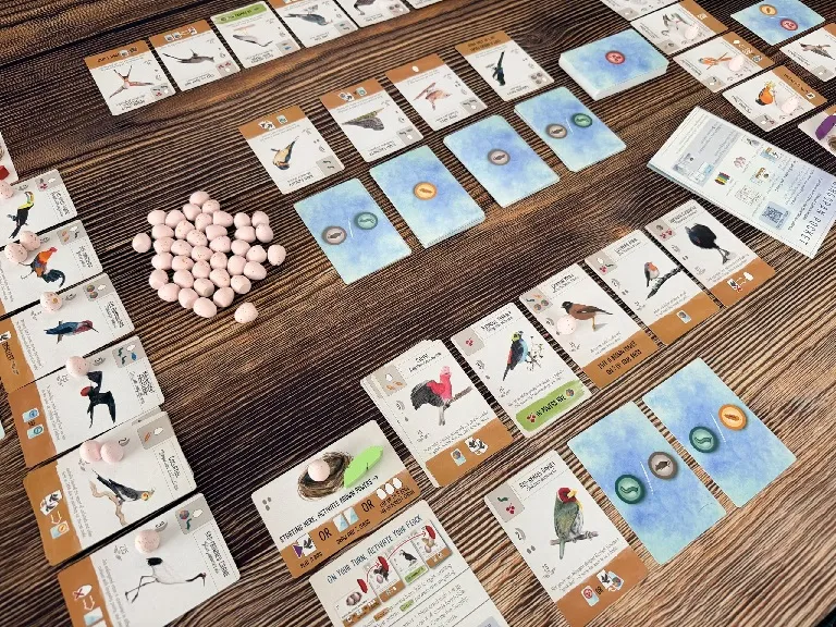 A wooden surface with a spread of cards from the Wingspan Pocket game. The cards display illustrations of various birds, eggs, food tokens, and nest images. Some cards have visible text such as "Starting Player: Activate Bird Powers", "Lay 1 Egg", "Gain 1 Food", and "Tuck 1 Card". There are also small, round food tokens in shades of green, orange, and white, and a pile of small, pink egg miniatures. The cards are arranged in a grid-like pattern, with some overlapping.