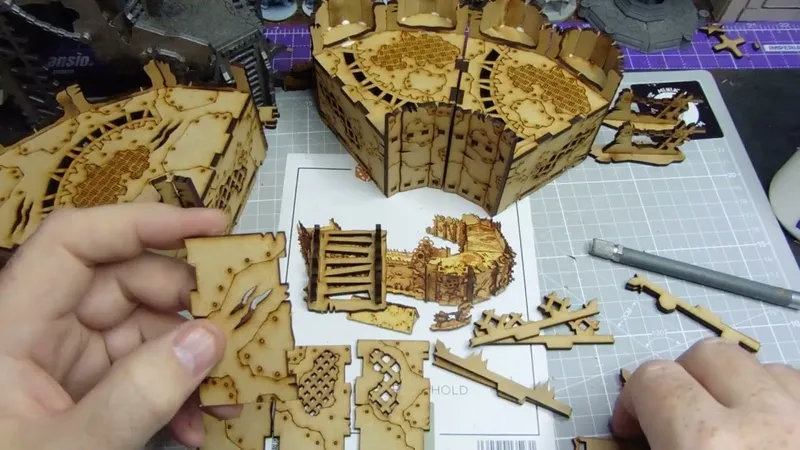 A hand holds a small, intricately cut piece of laser-cut wood, featuring detailed patterns and textures. Surrounding the hand are larger assembled sections of similar wooden terrain pieces, showcasing various designs, including grids and panels. In the background, a printed layout with additional terrain components is visible, indicating a crafting or gaming setup.