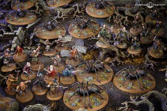 A detailed tabletop scene features various miniatures from the Middle-earth Strategy Battle Game, depicting a tense moment in Mirkwood. Prominent are several large spiders on circular bases, surrounded by a mix of characters including Bilbo, Thorin, and other dwarves, all positioned on a textured terrain with rocks and twisted roots. The dark, earthy colors of the setting enhance the atmosphere of the iconic "Flies and Spiders" chapter from The Hobbit.