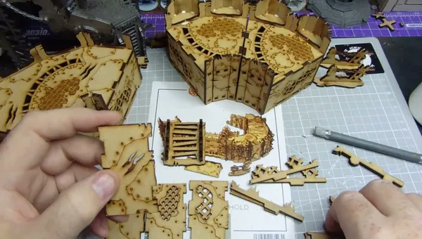 A hand holds a small, intricately cut piece of laser-cut wood, featuring detailed patterns and textures. Surrounding the hand are larger assembled sections of similar wooden terrain pieces, showcasing various designs, including grids and panels. In the background, a printed layout with additional terrain components is visible, indicating a crafting or gaming setup.
