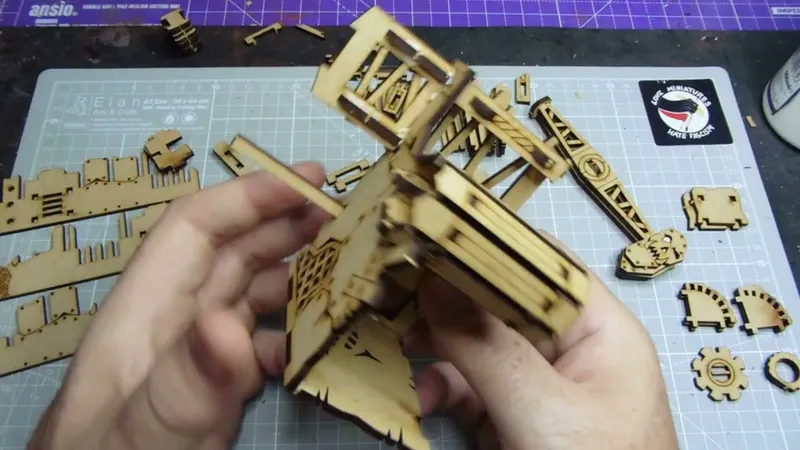 A partially assembled oil pump model made from laser-cut MDF is being held in hands, showcasing intricate details like vents and supports. Surrounding the model are various unassembled MDF pieces, indicating ongoing construction. A cutting mat with a grid pattern serves as the work surface, and a sticker reading "Orc Miniatures Hate Frogs" is visible in the background.