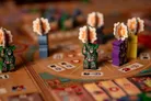 Three green devil miniatures with glowing maces stand on a board with a map of streets. Two drummers in blue and orange costumes are positioned behind them. Card tiles with icons and numbers, including one labeled "3D," are placed on the board.