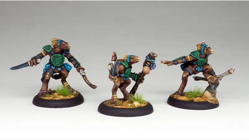 Three intricately painted miniatures stand on a textured base with tufts of grass. Each figure features a humanoid creature with a brown body, adorned with vibrant blue and green armor and accessories. The leftmost figure wields a curved sword, the center figure holds a staff with a small creature perched on top, and the rightmost figure points forward, showcasing their unique designs and expressions.