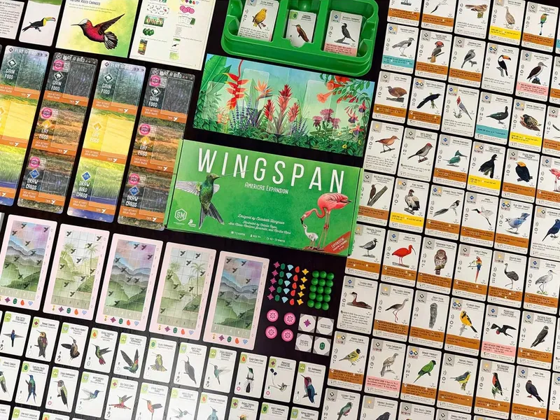 The image showcases the Wingspan: Americas Expansion board game, featuring a vibrant green box prominently displayed at the center, adorned with illustrations of a flamingo and a hummingbird. Surrounding the box are numerous bird cards, each depicting various species with detailed artwork and statistics. Additional components include goal cards, double-sided goal tiles, and colorful tokens, all set against a backdrop of lush tropical foliage.