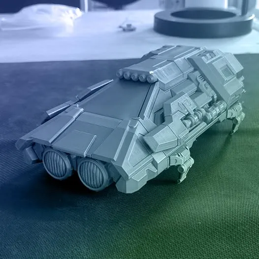 A detailed 3D-printed model of a futuristic vehicle, featuring a sleek, angular design with prominent headlights and various mechanical components. The model is displayed on a textured surface, with a blurred background that includes a circular object and some indistinct items. The overall color scheme is a monochromatic gray, highlighting the intricate details of the vehicle's structure.