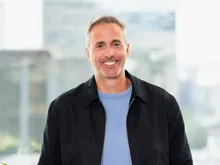A man in a black jacket over a blue shirt, smiling at the camera. Office setting with modern glass windows and indoor plants in the background.